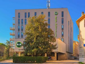 Modern beige building of B&B Hotel Fabriano Janus with multiple narrow windows, trees in front, and clear blue sky above