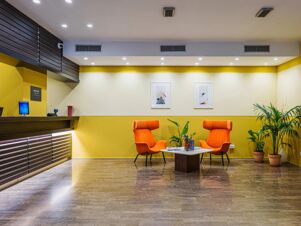 Reception area with a computer desk, two orange chairs, a small table with plants, and framed pictures at B&B Hotel Fabriano Janus