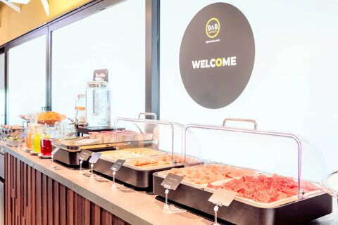Buffet counter with sliced meats, cheese, cereal, beverages, and a welcome sign at B&B Hotel Fabriano Janus