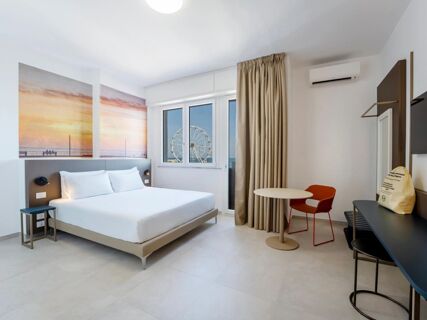 Modern hotel room at B&B Hotel Pesaro Des Bains featuring a double bed, wall art, window with a Ferris wheel view, curtains, table, and chair