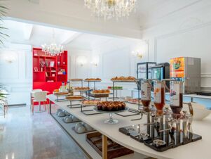 Buffet table with croissants, cakes, muffin, cereals, juice dispensers, crystal chandeliers, and red shelves in B&B Hotel Pesaro Des Bains