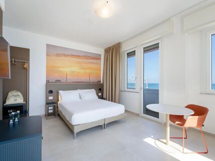 White bed with pillows under sunset artwork, window with Ferris wheel view, desk with chair in B&B Hotel Pesaro Des Bains