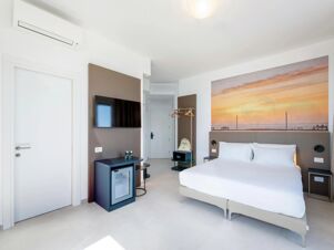 Bright hotel room at B&B Hotel Pesaro Des Bains with white bed, wall-mounted TV, mini fridge, air conditioner, and sunset artwork above bed