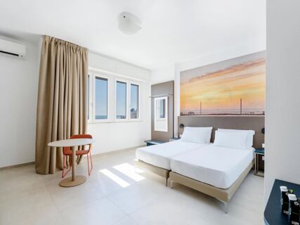 Bright hotel room at B&B Hotel Pesaro Des Bains with twin beds, large window, beige curtains, orange chair, and wall art of sunset sea view