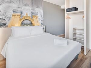 Double bed with white linens and pillows next to an open closet with hangers and a safe in a B&B Hotel Torino Orbassano room