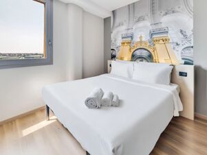 A double bed with white linens and rolled towels is set near a window in a room at B&B Hotel Torino Orbassano