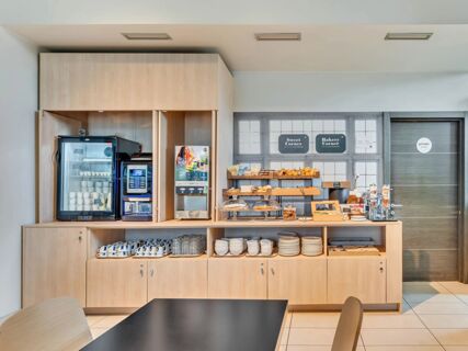 Breakfast area with coffee machines, refrigerated display, shelves with pastries, cups, plates, and a door labeled private at B&B Hotel Torino Orbassano