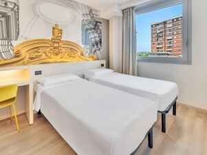Two single beds with white linens next to a window showing a building, a desk with yellow chair and decorative wall art at B&B Hotel Torino Orbassano