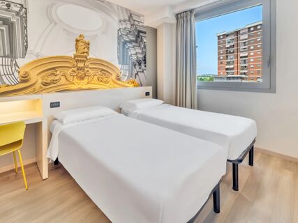 Two single beds with white linens next to a window showing a building, a desk with yellow chair and decorative wall art at B&B Hotel Torino Orbassano