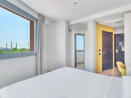 Bright hotel room at B&B Hotel Torino Orbassano with a window view, white walls, a full-length mirror, and a yellow chair