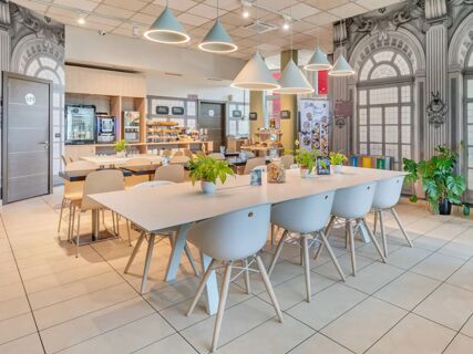 Bright dining area with white tables and chairs, hanging cone lights, plants, and decorative wall murals at B&B Hotel Torino Orbassano