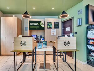 Reception area at B&B Hotel Torino Orbassano with two counters, wooden panels, high stools, green ceiling, and snack rack on the side