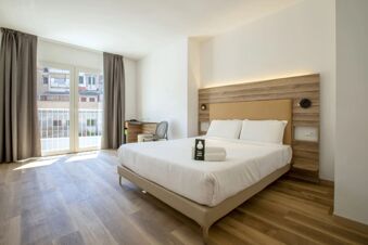 B&B Hotel Sassari room with a large bed, white linens, wooden headboard, bedside lights, balcony door with gray curtains, and desk with chair