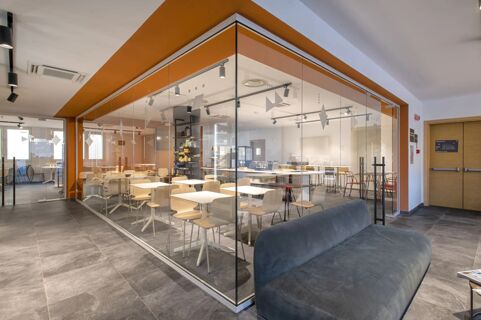 View of a modern dining area with wooden chairs and white tables behind glass walls inside B&B Hotel Sassari lobby space
