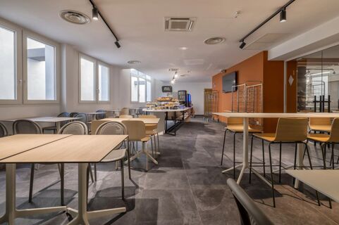 Spacious dining area at B&B Hotel Sassari with multiple tables and chairs, large windows, and a buffet station with baked goods and a coffee machine