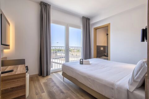 Bright hotel room at B&B Hotel Sassari with a neatly made bed, wooden desk, balcony doors with gray curtains, and bathroom door visible