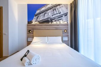 Double bed with beige headboard, two rolled white towels, dark gray curtains, and building photo above bed at B&B Hotel Sassari