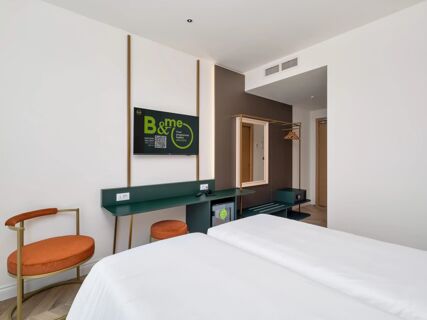 A hotel room at B&B Hotel Catania Bellini with a green desk, brown chair and stool, a wall-mounted TV, and a mirror with coat hangers nearby