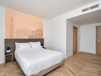 White bed with pillows next to wall art of buildings, wooden doors, and wood pattern floor at B&B Hotel Catania Bellini