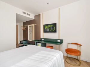 White bedspread on bed with green desk, orange chair, wall-mounted TV, mirror, and coat rack in B&B Hotel Catania Bellini room