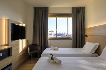 Two single beds with white linens and towels face a window with curtains and a mounted TV at B&B Hotel Catania City Center