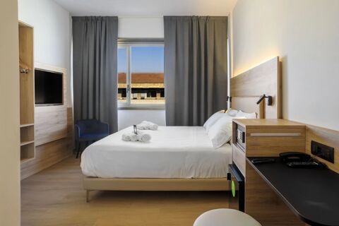 A modern hotel room at B&B Hotel Catania City Center with a double bed, white linens, folded towels, TV, desk, and window with curtains