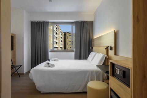 Double bed with white linens and pillows, bedside reading lights, a window with gray curtains, and a safe in the room at B&B Hotel Catania City Center