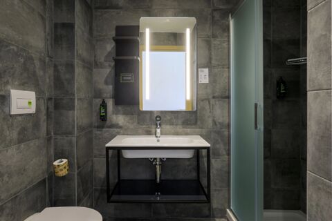 Modern bathroom interior at B&B Hotel Catania City Center with gray tiles, illuminated mirror, white sink, and frosted glass shower door