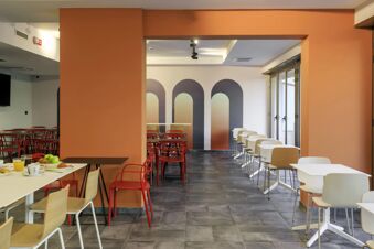 Dining area at B&B Hotel Catania City Center with red and wooden chairs, white tables, orange walls, and breakfast items on a table