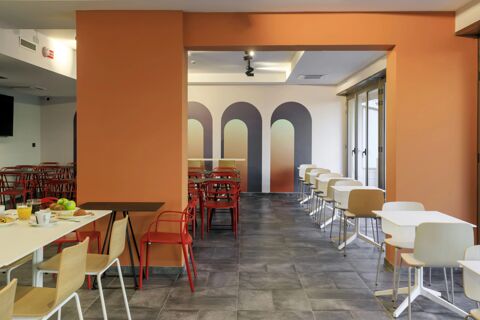 Dining area at B&B Hotel Catania City Center with red and wooden chairs, white tables, orange walls, and breakfast items on a table
