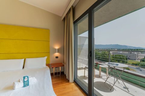 Room at B&B Hotel Pisa Airport with white bed linens, yellow headboard, bedside table, sliding doors, and a balcony with table and stools