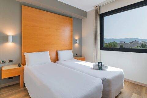 Two neatly made single beds with white linens, wooden headboard, bedside tables, and a window showing a cityscape at B&B Hotel Pisa Airport