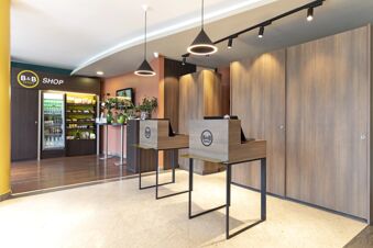 Reception area with two wooden desks, pendant lights, plants on tables, and a snack shop at B&B Hotel Pisa Airport