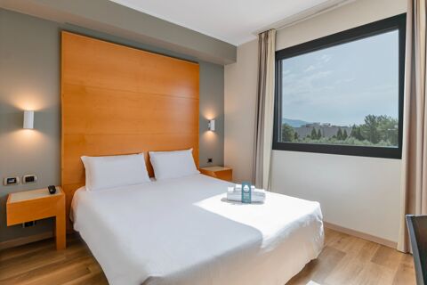 Double bed with white linens and pillows in a room at B&B Hotel Pisa Airport with wooden accents and a large window showing trees outside