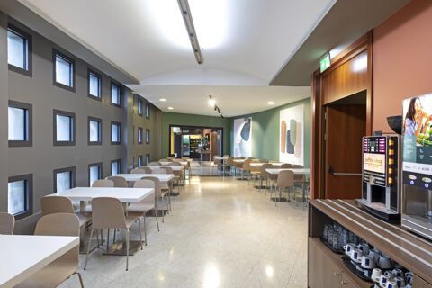 Dining area with white tables and wooden chairs, vending machines, green and gray walls, at B&B Hotel Pisa Airport