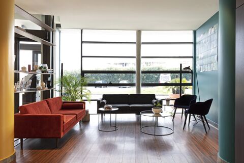 Lounge area at B&B Hotel Pisa Airport with large windows, a red sofa, black sofa, blue chairs, tables, and decorative plants