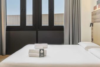 White bed with folded towels, window with black frames, beige headboard, wall lamp, and curtains in B&B Hotel Prato City Center