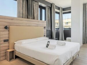 Bright hotel room at B&B Hotel Prato City Center with a double bed, wooden headboard, folded towels, and gray curtains on windows