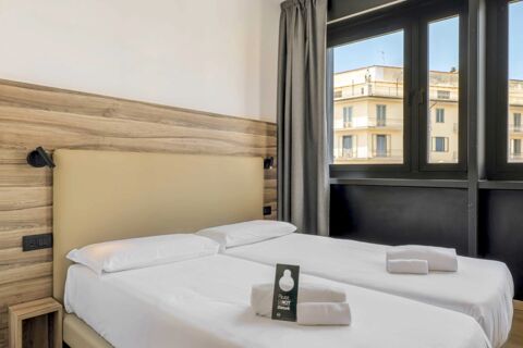 Twin beds with white linens and pillows beside a window showing surrounding buildings at B&B Hotel Prato City Center