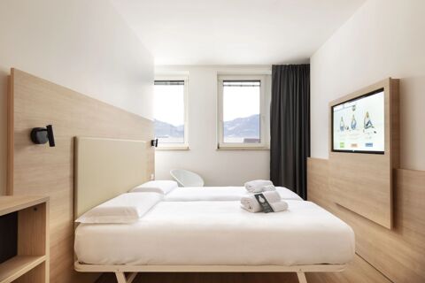Two single beds with white linens and folded towels in a bright room with two windows and a mounted TV at B&B Hotel Bolzano