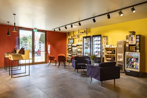 Lobby area of B&B Hotel Bolzano with red and yellow walls, vending machines, blue and orange chairs, and glass door exit