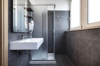 Modern bathroom with glass shower enclosure, white sink, wall-mounted hairdryer, and dark gray tiles at B&B Hotel Bolzano