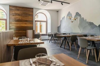 Dining area with wooden tables, grey chairs, large arched windows, mountain mural, and bird decorations in B&B Hotel Passo Tre Croci Cortina