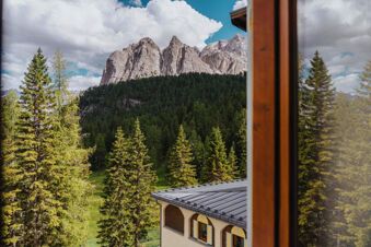 View of rocky mountains and pine trees from a window with part of the roof and facade of B&B Hotel Passo Tre Croci Cortina visible