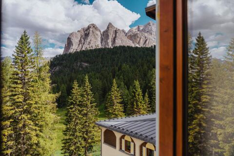 View of rocky mountains and pine trees from a window with part of the roof and facade of B&B Hotel Passo Tre Croci Cortina visible
