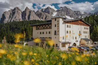 B&B Hotel Passo Tre Croci Cortina building with white walls and wooden accents surrounded by green forest and rocky mountains in background