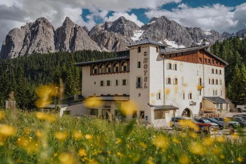 B&B Hotel Passo Tre Croci Cortina building with white walls and wooden accents surrounded by green forest and rocky mountains in background
