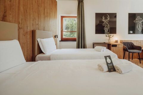 Two single beds with white linens and pillows, a wooden nightstand with a lamp, chair, window, and modern deer artwork in a B&B Hotel Passo Tre Croci Cortina room