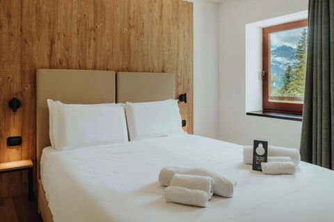 A double bed with white linens and pillows against a wooden headboard and a window showing a mountain view at B&B Hotel Passo Tre Croci Cortina