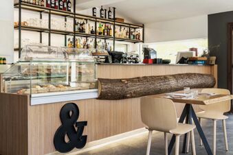 Modern bar counter with glass display of pastries, bottles on shelves, coffee machine, and wooden tables at B&B Hotel Passo Tre Croci Cortina